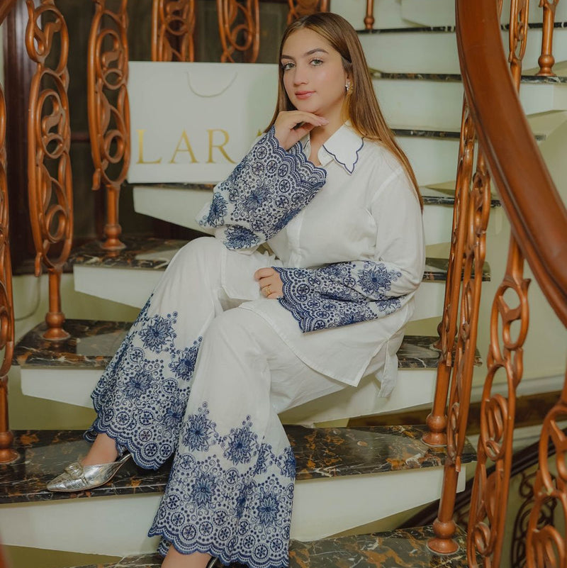 Woman sitting on a staircase wearing a white outfit with blue floral patterns, with a 'LAR' sign in the background.